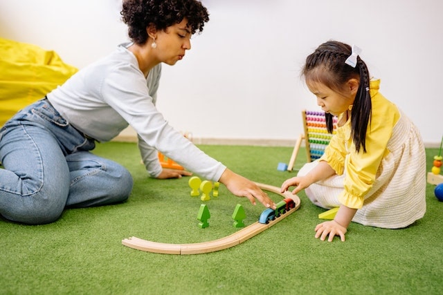 Image of a preschool teacher sitting on the floor and playing with a young child, engaging in a fun and educational activity together in a classroom.
