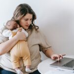 A stressed mother multitasking, working on a laptop while attending to her child, illustrating the challenges and signs of Depleted Mom Syndrome. 