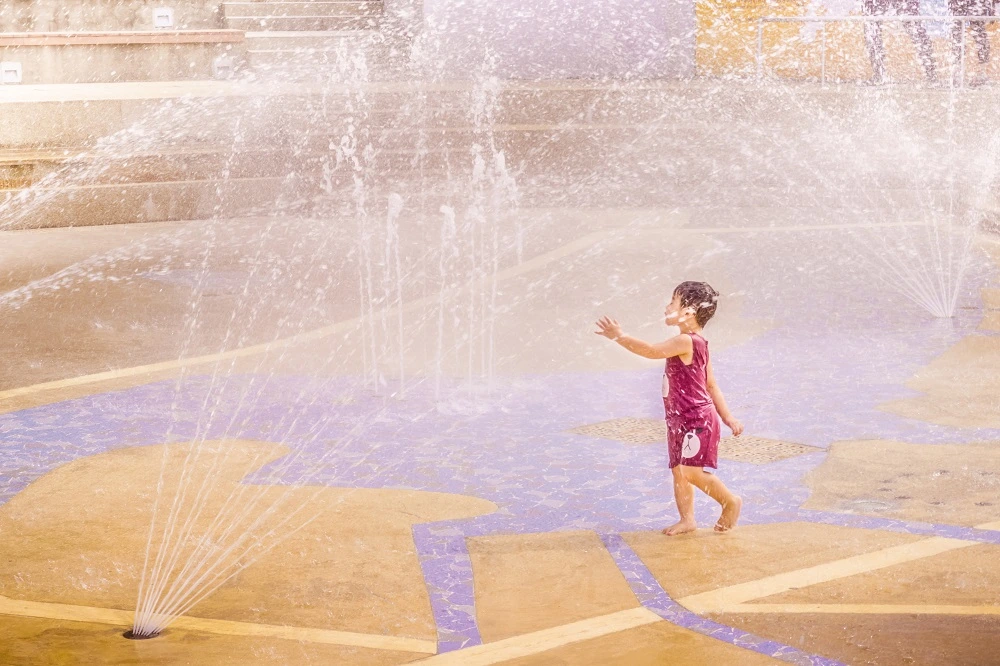 Child playing joyfully in a water fountain splash area at a park, enjoying the refreshing water jets.