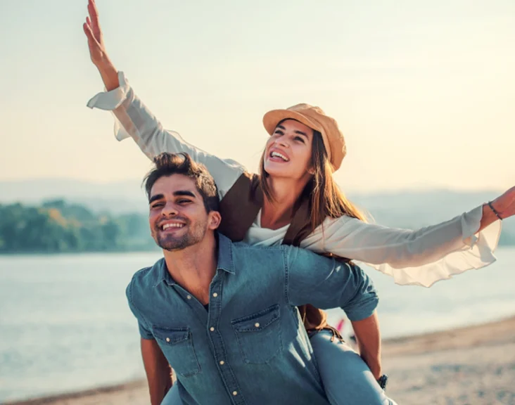 Understanding Complacency in Relationships. Joyful couple by the lake, with the woman playfully riding on the man's back, enjoying a carefree moment together.