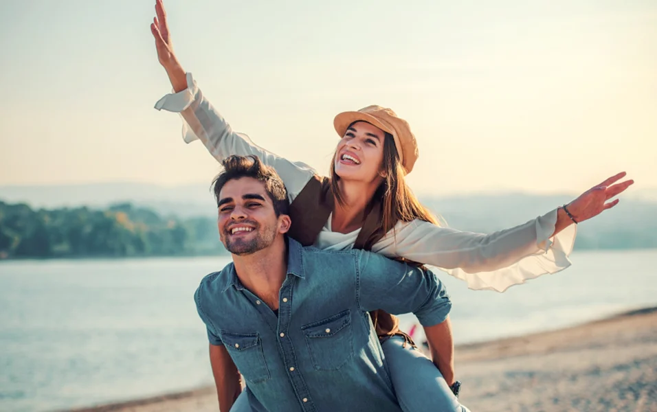 Understanding Complacency in Relationships. Joyful couple by the lake, with the woman playfully riding on the man's back, enjoying a carefree moment together.