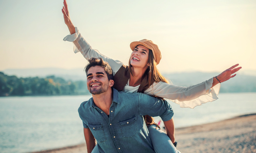 Understanding Complacency in Relationships. Joyful couple by the lake, with the woman playfully riding on the man's back, enjoying a carefree moment together.